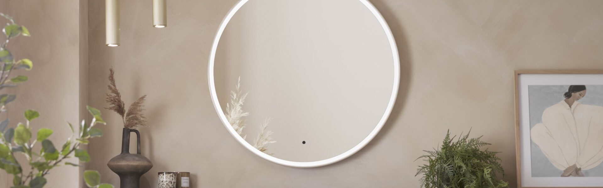 A round, illuminated mirror with a brass frame. It is hanging on a wall in a modern bathroom setting. There are also two gold pendant lights, a vase with dried flowers, a framed picture, and a potted plant.