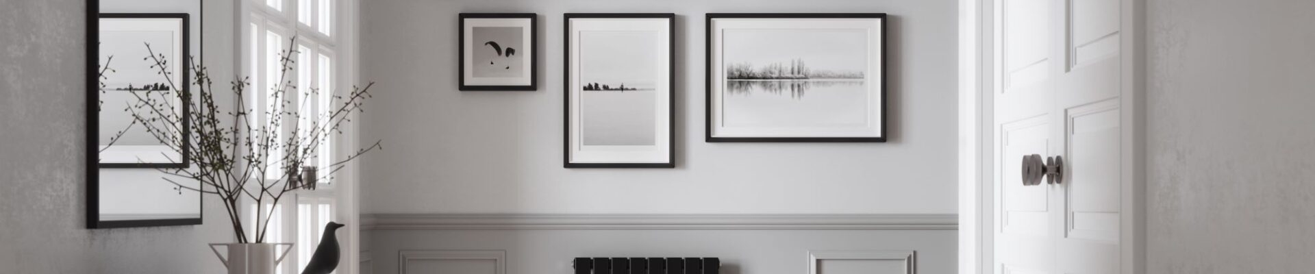 The images shows a single panel horizontal radiator with a matte black finish. It is positioned below a gallery wall of framed photographs. The hallway has grey walls, a wooden floor, and a door leading to another room.