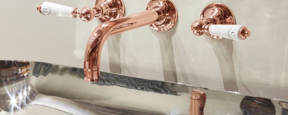 A luxurious bathroom vanity with a copper-coloured tap set. The tap has a classic design with white porcelain handles. The basin is rectangular and made of polished stainless steel. The overall aesthetic of the bathroom is elegant and sophisticated.