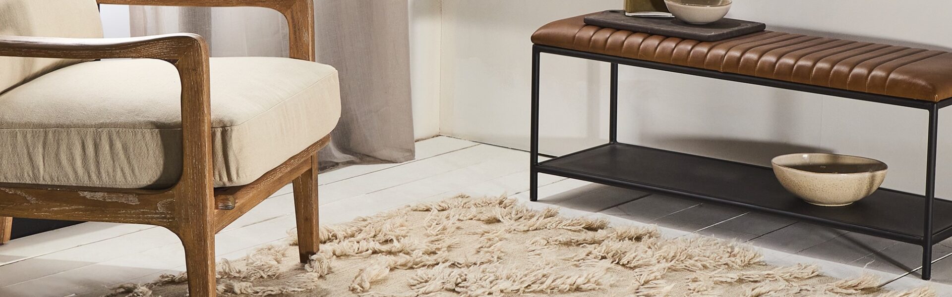 A living room with a beige rug, a wooden armchair, a metal bench with a leather seat, and a vase with dried flowers. The overall style of the room is bohemian and cosy.