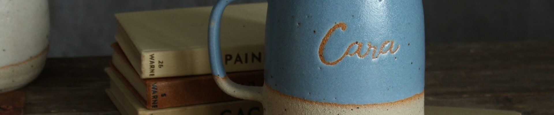 The image conveys a sense of warmth and personalisation. The mug feels like a cherished object with the name "Cara" etched into it, and the books suggest a cosy, personal space.