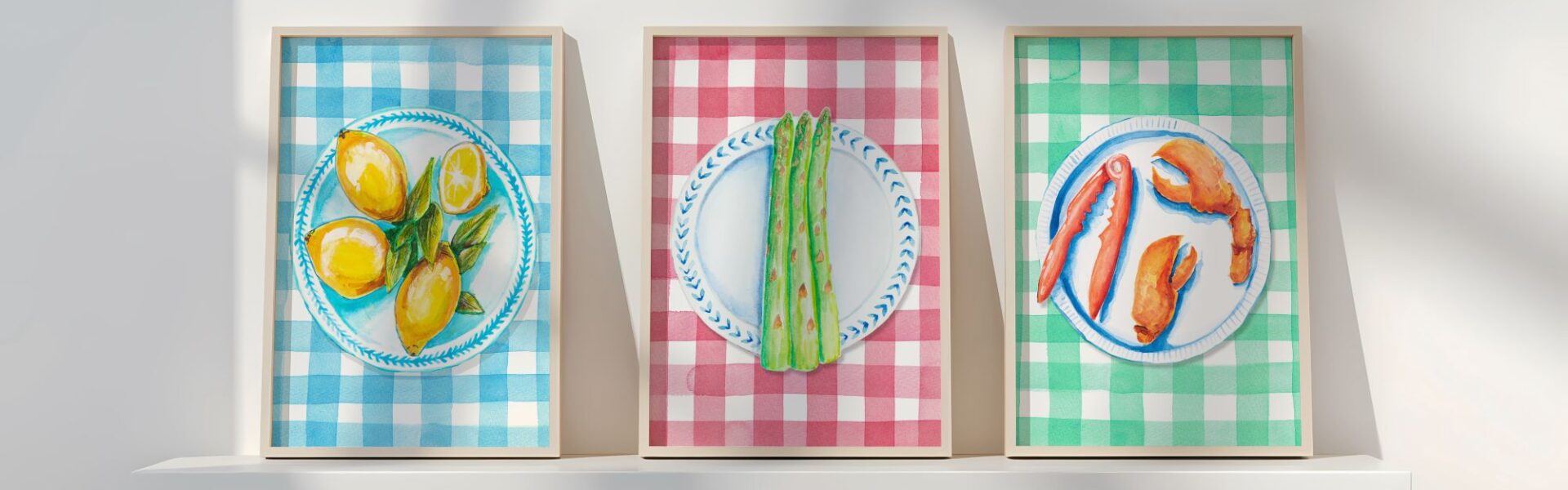 Three framed illustrations of food on plates. The left image shows lemons on a blue and white plate against a blue and white gingham background. The middle image shows asparagus on a blue and white plate against a red and white gingham background. The right image shows lobster claws on a blue and white plate against a green and white gingham background.