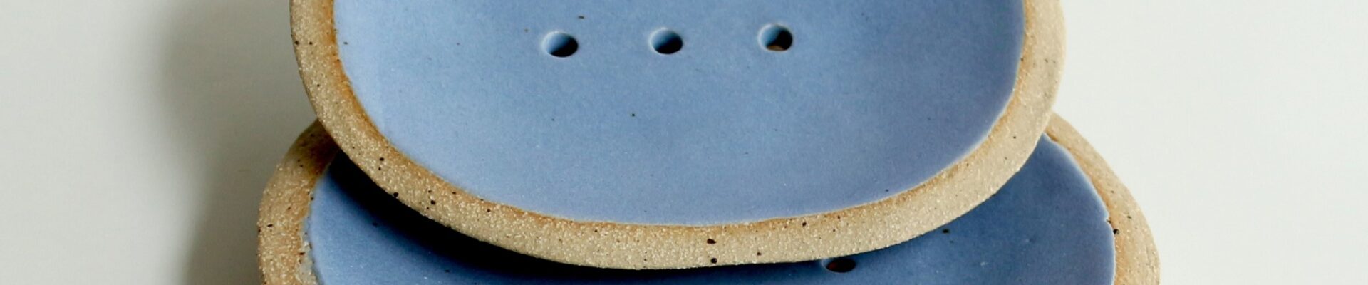 Two blue ceramic soap dishes with a raised rim and drainage holes. The soap dishes are slightly textured and have a natural, organic look.