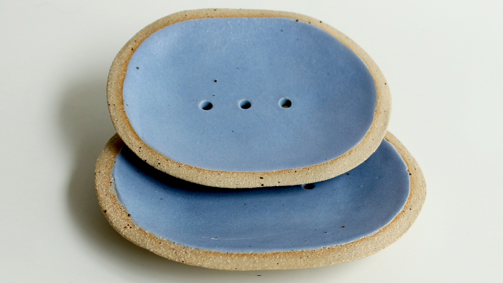 Two blue ceramic soap dishes with a raised rim and drainage holes. The soap dishes are slightly textured and have a natural, organic look.