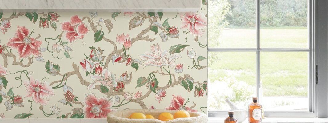 A bright kitchen featuring Ronald Redding Classics Marguerite Vine wallpaper in a pink and green botanical pattern, white marble countertops, a vintage stone bowl filled with oranges, and two bottles on the counter.