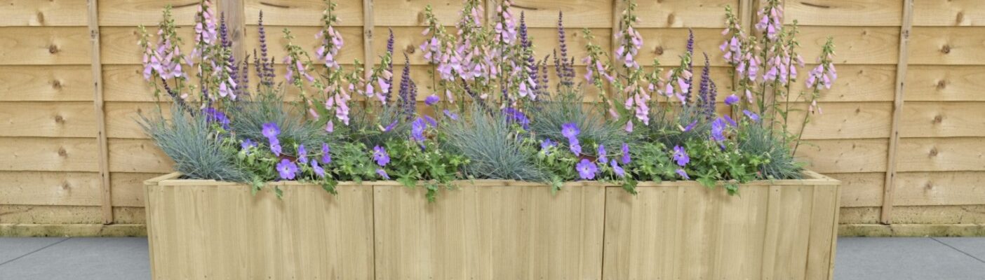 A rectangular wooden planter box sits on a grey tiled surface against a light wood fence. The planter is filled with a variety of purple and lavender flowers.