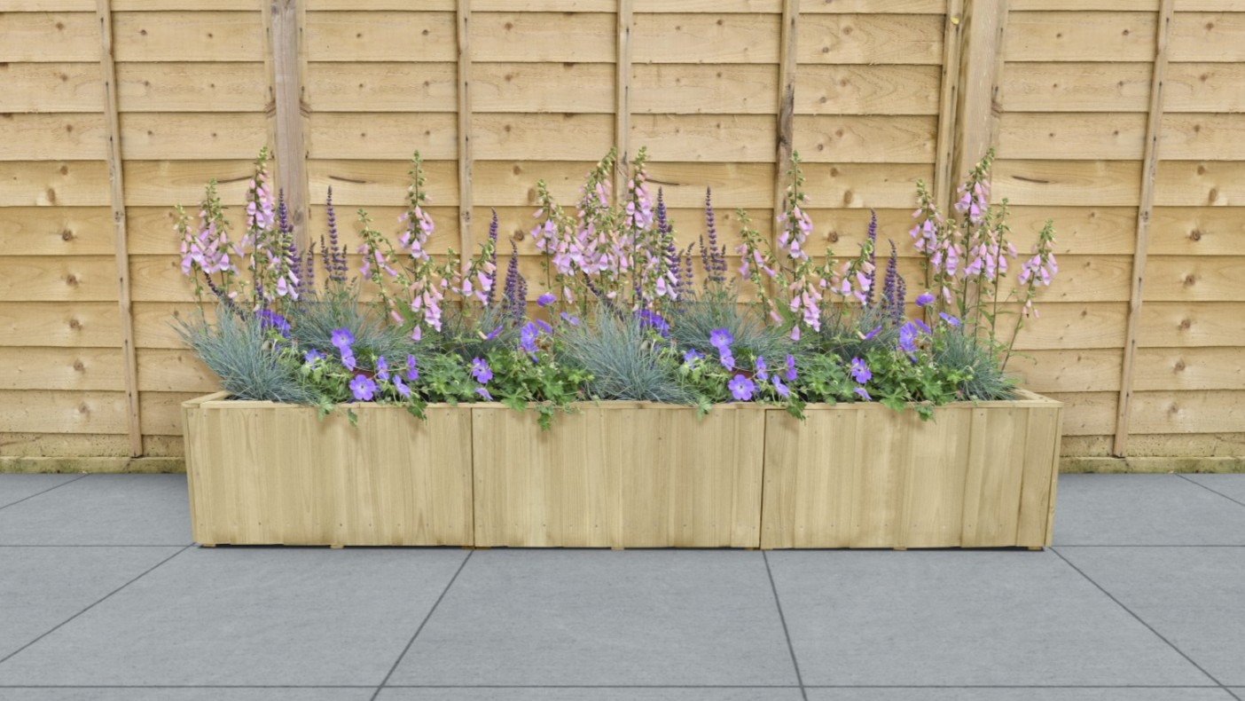 A rectangular wooden planter box sits on a grey tiled surface against a light wood fence. The planter is filled with a variety of purple and lavender flowers.