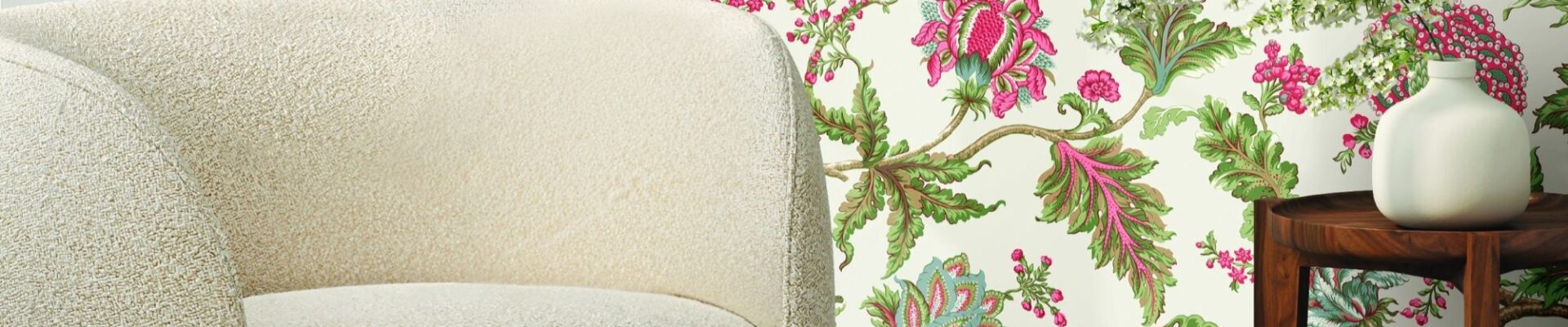 A close-up shot of a cream-coloured, rounded boucle fabric chair with a dark wood base, positioned in front of a wall with a vibrant floral wallpaper featuring pink, green, and white flowers and leaves.