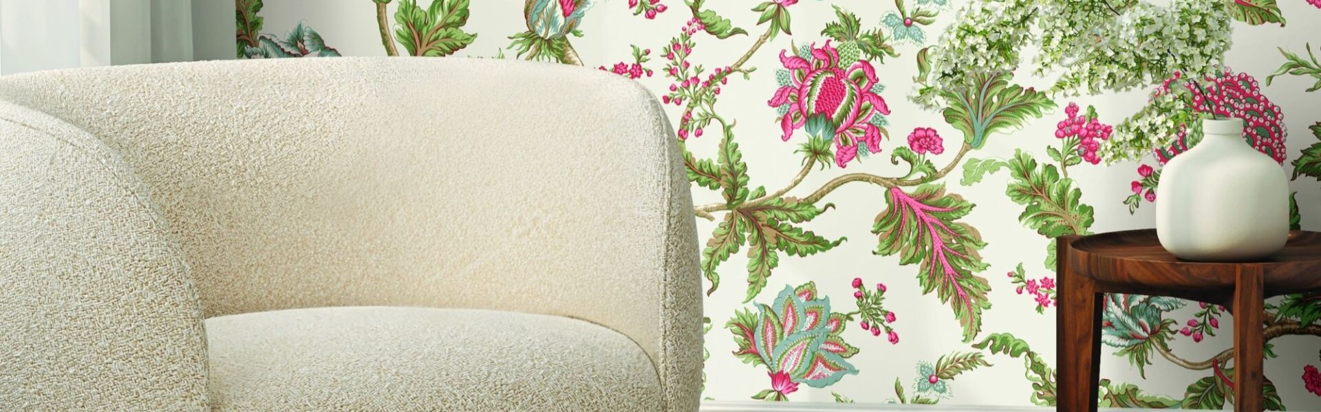 A close-up shot of a cream-coloured, rounded boucle fabric chair with a dark wood base, positioned in front of a wall with a vibrant floral wallpaper featuring pink, green, and white flowers and leaves.