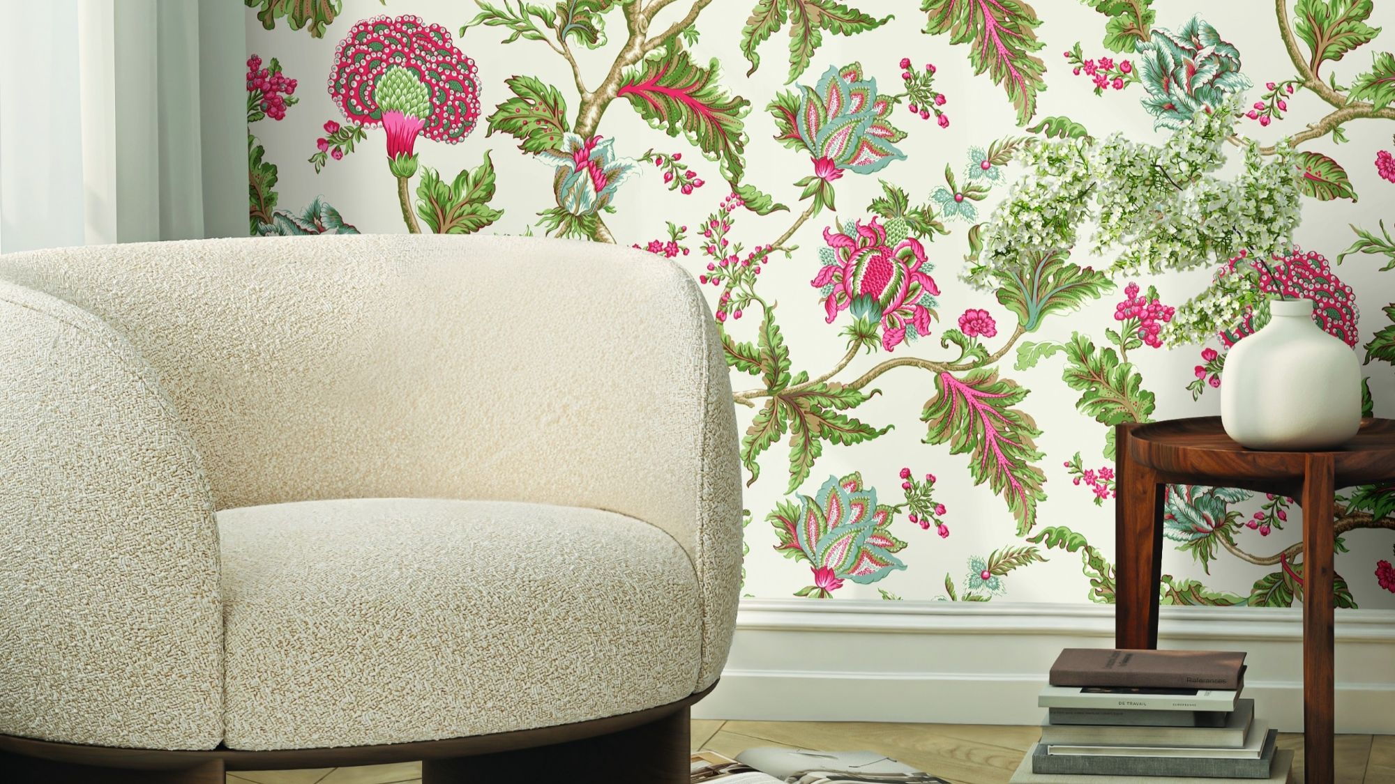 A close-up shot of a cream-coloured, rounded boucle fabric chair with a dark wood base, positioned in front of a wall with a vibrant floral wallpaper featuring pink, green, and white flowers and leaves.