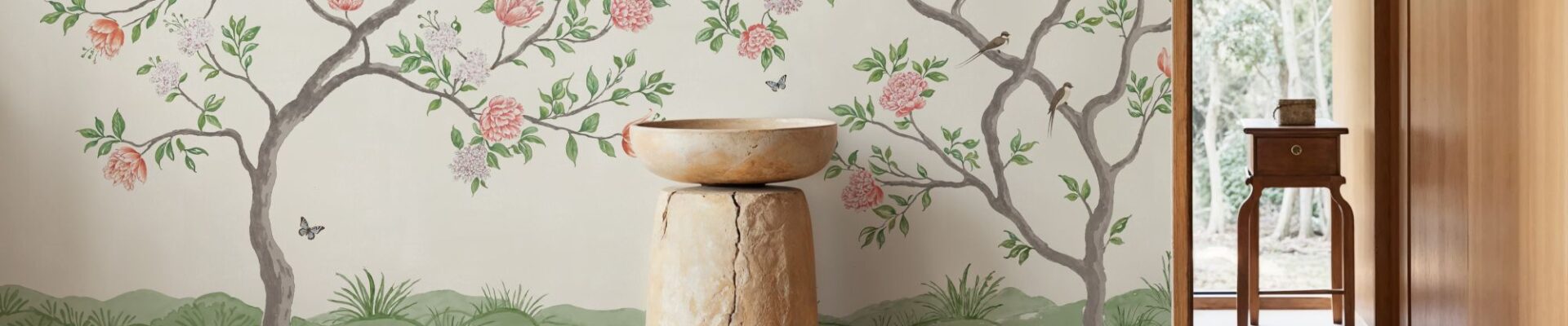 A bright, indoor space featuring a mural-style wallpaper of flowering trees against a light background. A tall, rustic pedestal made of wood and stone holds a simple bowl in the foreground. The room has light wood floors and a doorway leading to an outdoor space with lush greenery.