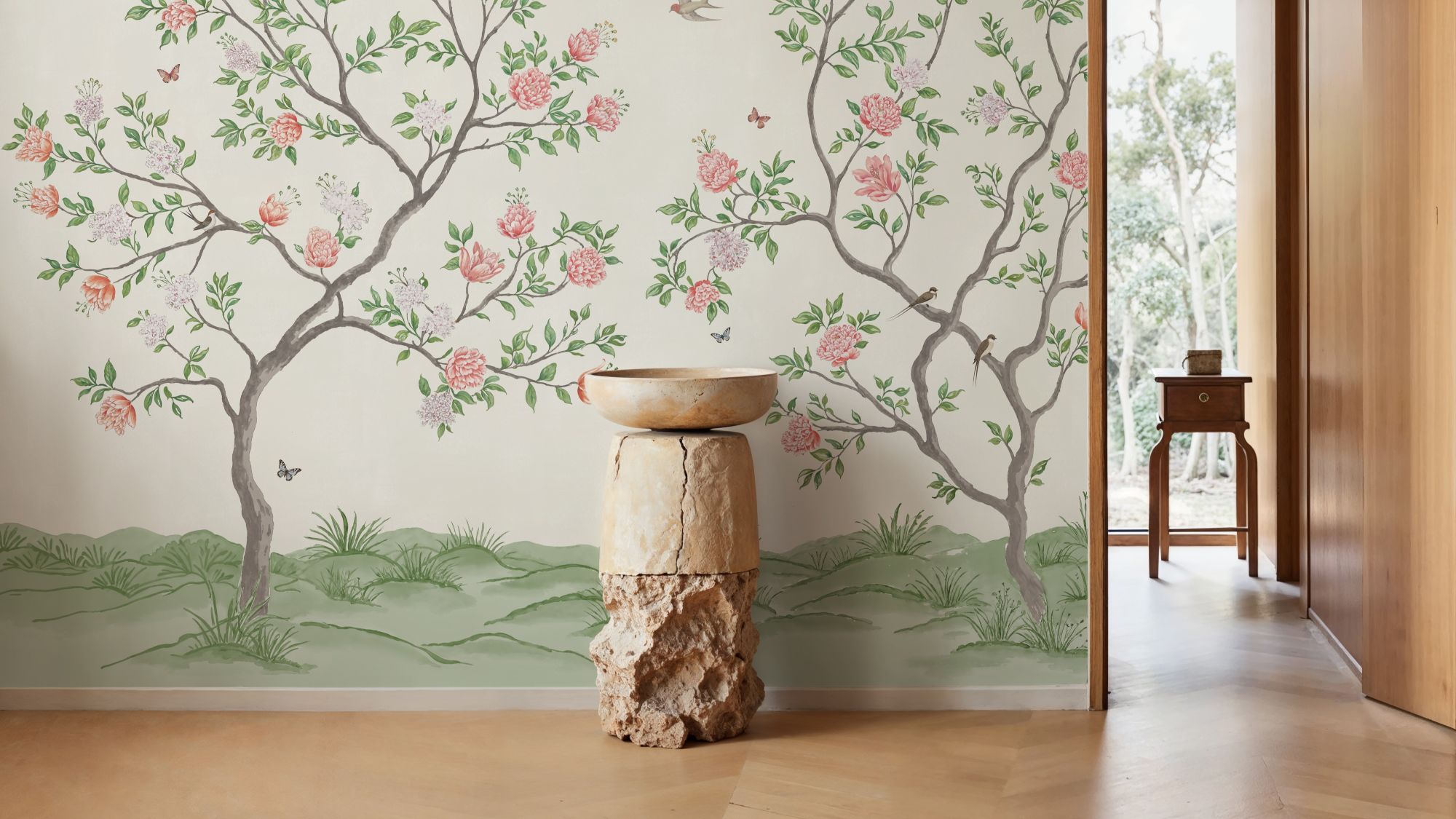 A bright, indoor space featuring a mural-style wallpaper of flowering trees against a light background. A tall, rustic pedestal made of wood and stone holds a simple bowl in the foreground. The room has light wood floors and a doorway leading to an outdoor space with lush greenery.