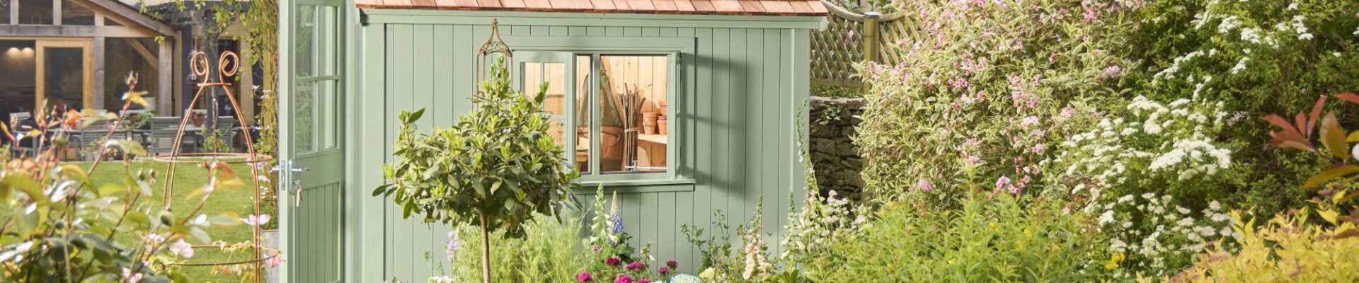 The primary focus is the potting shed, painted in a soft, muted green. It has a traditional design with vertical plank siding and a pitched roof covered in small, rectangular cedar shingles. The double doors have simple panelled detailing.