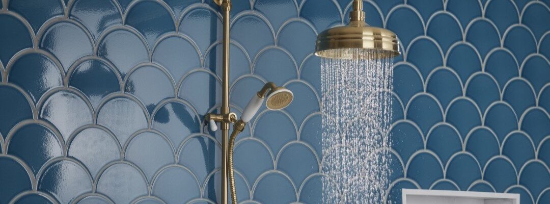 A close-up shot of a brass-finished shower system against a backdrop of blue, fish-scale patterned tiles. Water streams from the overhead rain shower head. A handheld shower wand is also visible on the left.