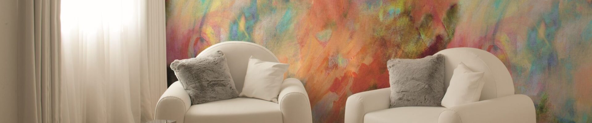 A bright, modern room features two white armchairs facing each other, with a vibrant, abstract painting-like mural on the wall behind them. The mural is a mix of swirling colours including yellow, orange, red, purple, blue, and green.