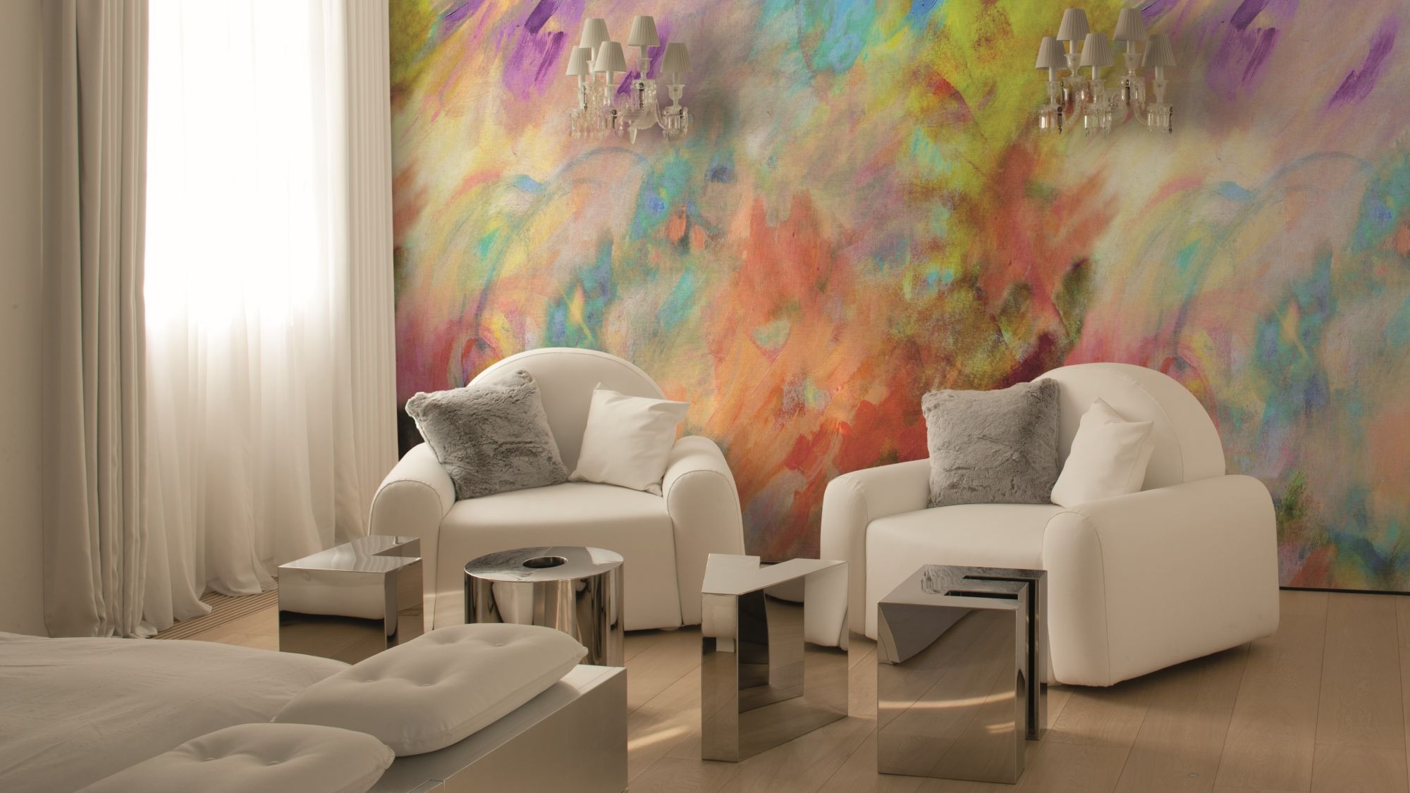 A bright, modern room features two white armchairs facing each other, with a vibrant, abstract painting-like mural on the wall behind them. The mural is a mix of swirling colours including yellow, orange, red, purple, blue, and green.