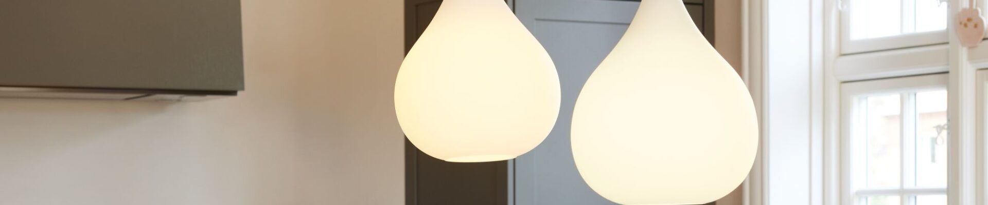 A close-up interior shot shows two teardrop-shaped pendant lights hanging in a kitchen. The lights are made of white, frosted glass and emit a soft, warm glow. They hang from thin black cords and have a small metallic accent at the top.