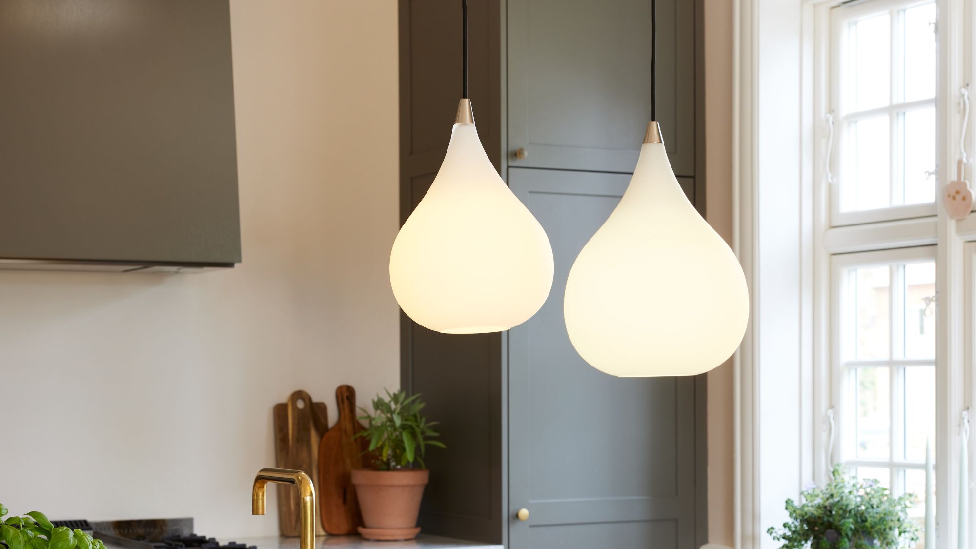 A close-up interior shot shows two teardrop-shaped pendant lights hanging in a kitchen. The lights are made of white, frosted glass and emit a soft, warm glow. They hang from thin black cords and have a small metallic accent at the top.