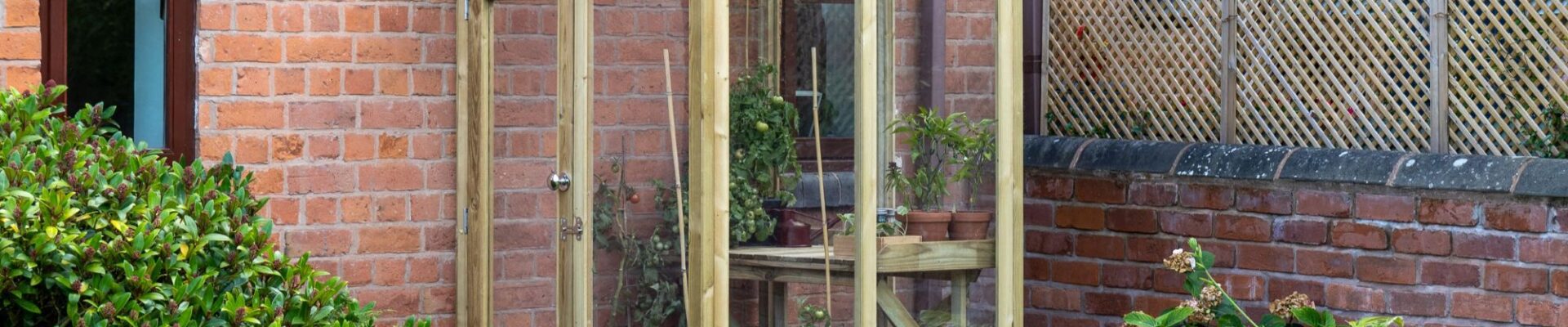 Outdoor image of a wooden lean-to greenhouse with a single door and clear panels, positioned against a brick wall with arched windows. Potted plants are visible inside.