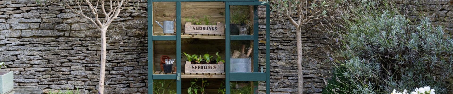 A dark green, A-frame greenhouse with wooden construction and glass panels sits in a garden setting.
