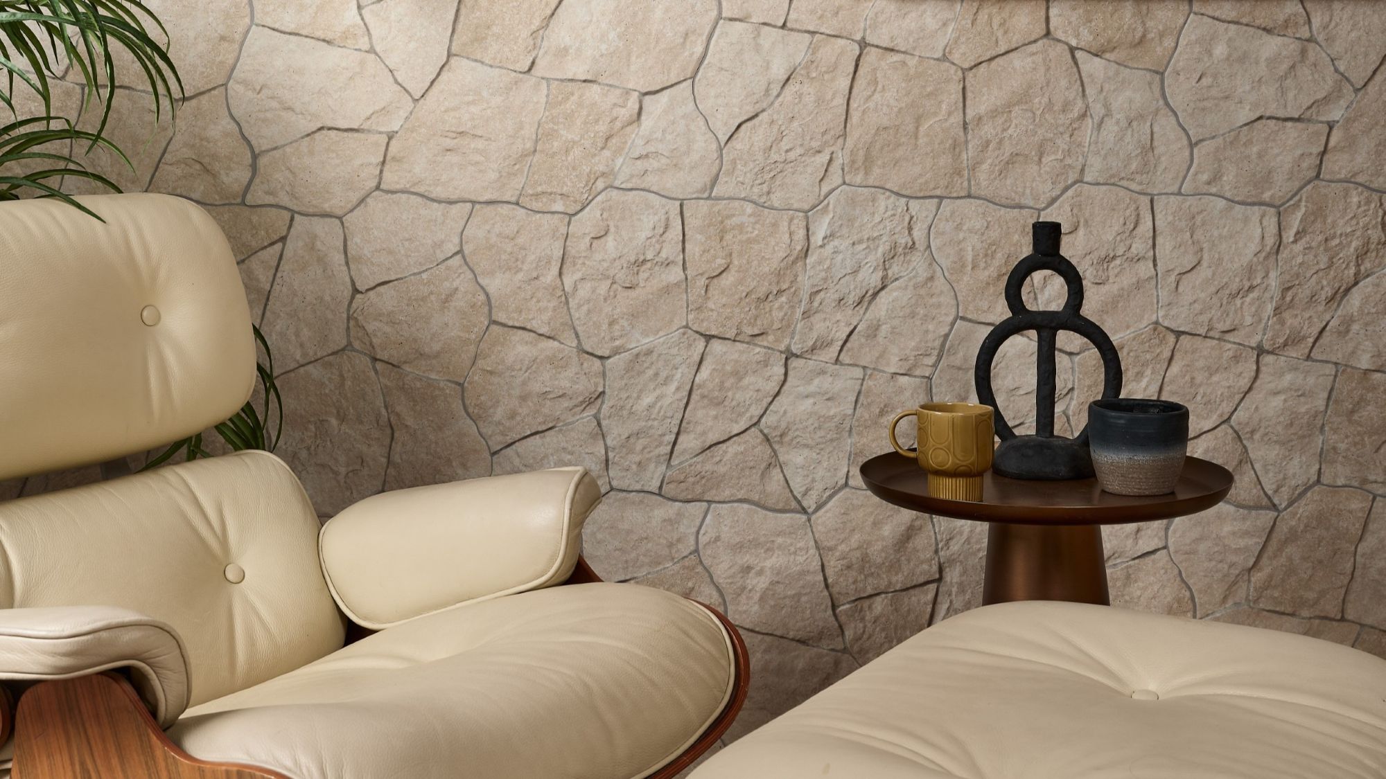 A modern living room corner featuring a cream-coloured leather armchair with a matching ottoman, positioned in front of a textured stone wall