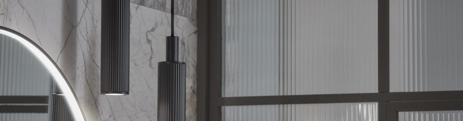 The primary focus is two pendant lights, which are identical in design. They are long, cylindrical, and appear to be made of a dark-coloured metal, possibly with a textured or ribbed surface. They hang from thin cables, suggesting a minimalist and contemporary style. The lights are positioned at slightly different heights, adding visual interest.