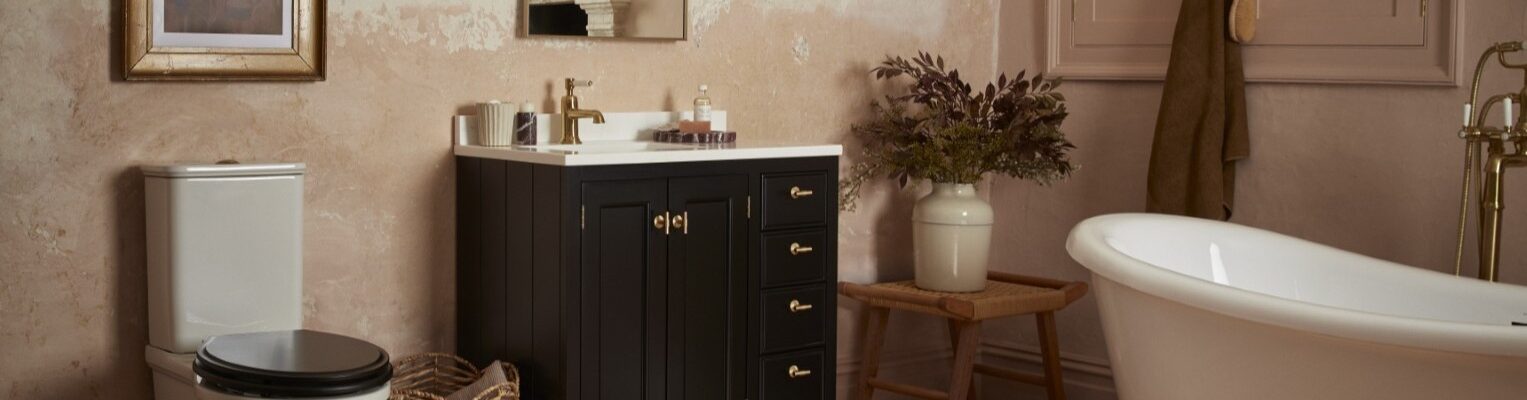 A bathroom with a rustic, vintage aesthetic. The walls have a textured, plaster-like finish in a light pinkish-beige hue. A dark wood vanity with a white countertop and gold hardware is centred in the frame
