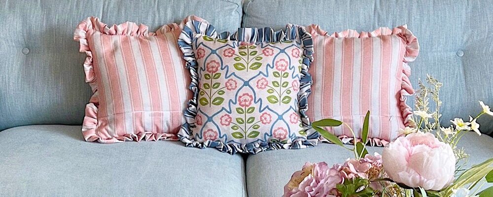 A close-up shot of a light blue tufted couch with three decorative pillows. The two outer pillows feature pink and white vertical stripes with ruffled edges. The centre pillow has a floral pattern with pink flowers and blue accents on a white background, also with ruffled edges.