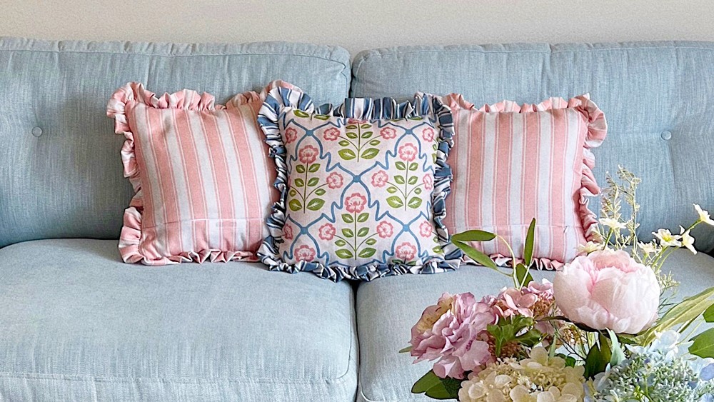A close-up shot of a light blue tufted couch with three decorative pillows. The two outer pillows feature pink and white vertical stripes with ruffled edges. The centre pillow has a floral pattern with pink flowers and blue accents on a white background, also with ruffled edges.