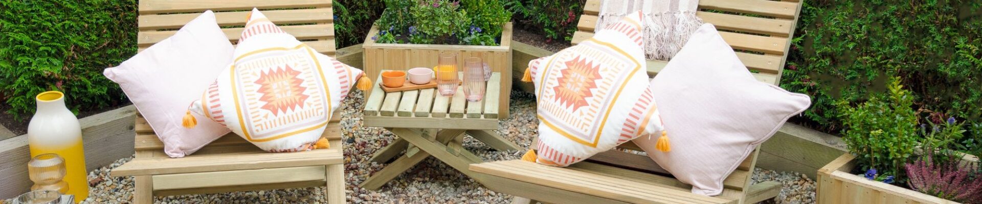 Two slatted wooden chairs and a small slatted wooden table arranged on a gravel surface. The chairs have cushions on them, and there are a few decorative items on the table, along with lanterns on the ground nearby.