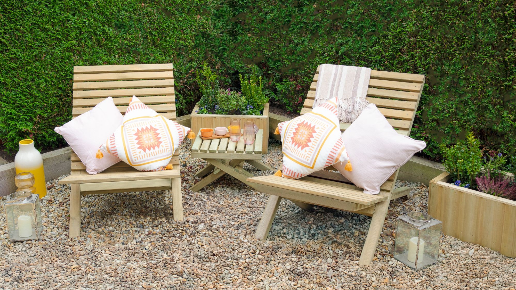 Two slatted wooden chairs and a small slatted wooden table arranged on a gravel surface. The chairs have cushions on them, and there are a few decorative items on the table, along with lanterns on the ground nearby.