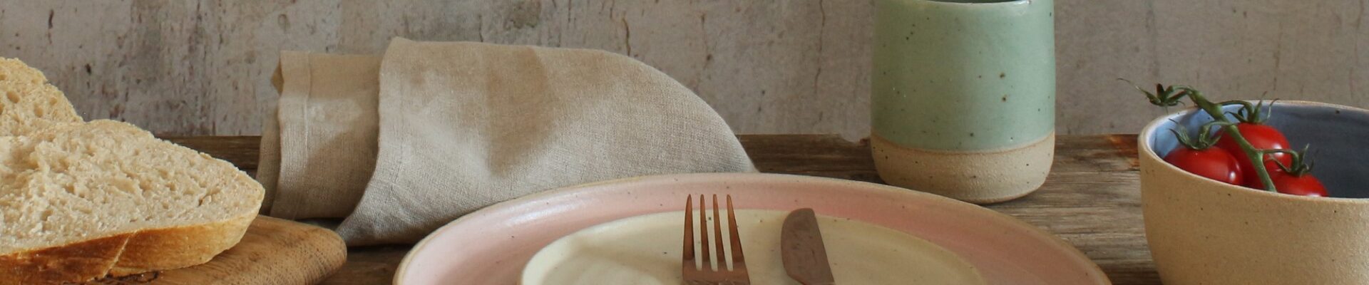 A stack of two ceramic plates. The larger plate has a light beige exterior and a soft pink interior. The smaller plate is a creamy white. A fork and knife rest on the smaller plate.