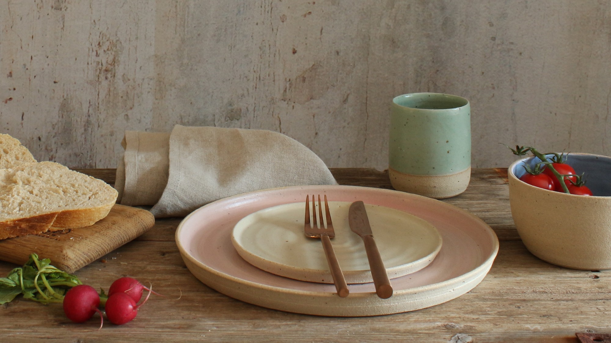 A stack of two ceramic plates. The larger plate has a light beige exterior and a soft pink interior. The smaller plate is a creamy white. A fork and knife rest on the smaller plate.