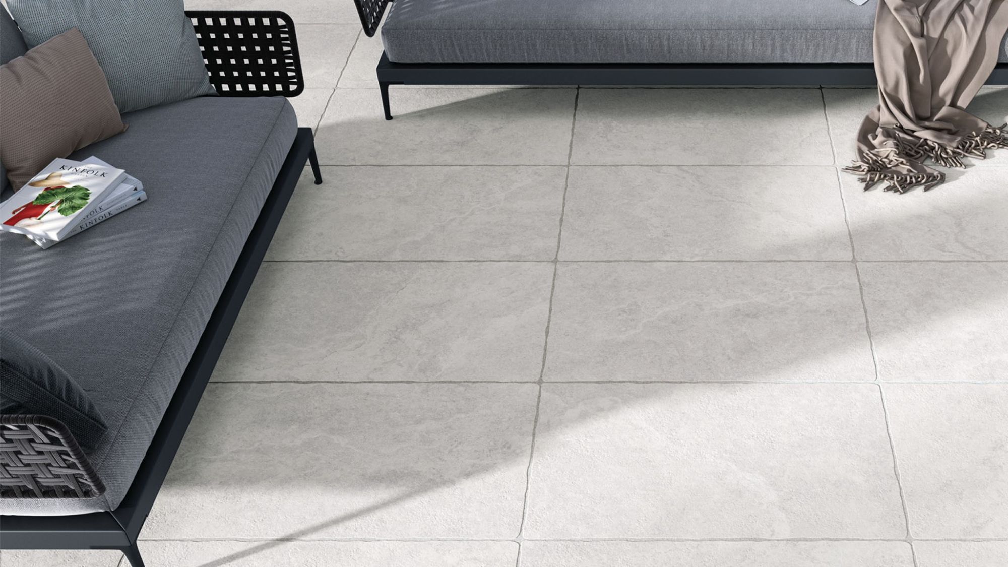 A top-down view shows a section of a patio or outdoor space paved with large, light grey rectangular tiles. Two modern outdoor sofas with grey cushions and black frames are partially visible on the left and top edges.