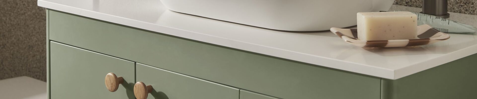 The image shows a close-up of a bathroom vanity unit. The vanity has a modern, minimalist design with a light sage green cabinet and a white countertop. A sleek, white, oval-shaped countertop basin sits on top of the counter.