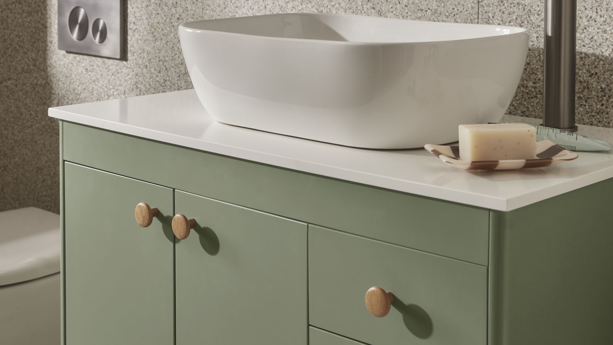 The image shows a close-up of a bathroom vanity unit. The vanity has a modern, minimalist design with a light sage green cabinet and a white countertop. A sleek, white, oval-shaped countertop basin sits on top of the counter.
