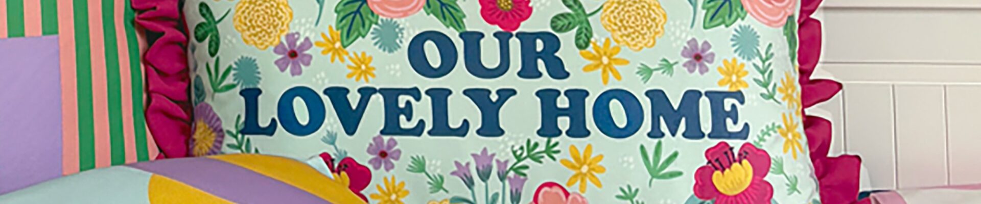 Close-up of a decorative pillow on a bed featuring a floral design in pink, yellow, and green on a light blue background, with the phrase ‘Our Lovely Home’ prominently displayed
