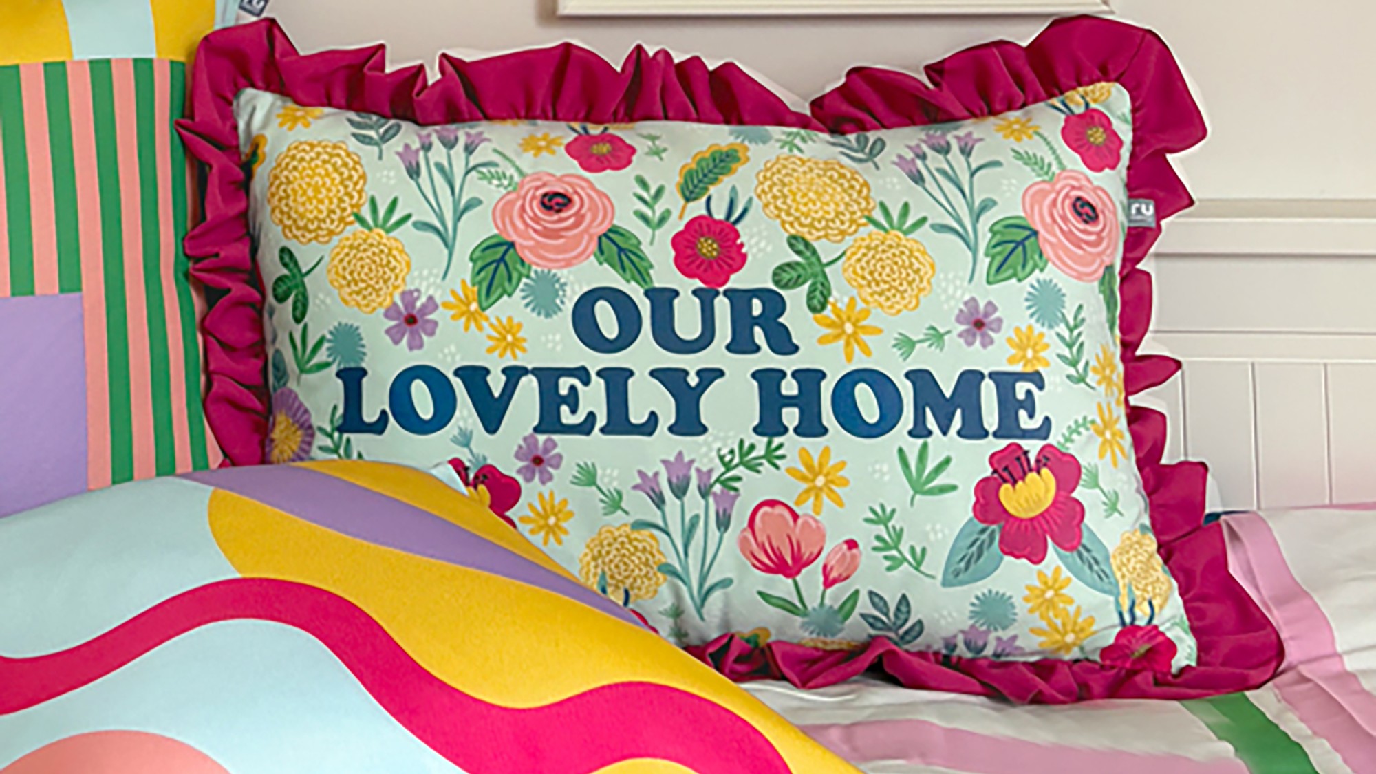 Close-up of a decorative pillow on a bed featuring a floral design in pink, yellow, and green on a light blue background, with the phrase ‘Our Lovely Home’ prominently displayed