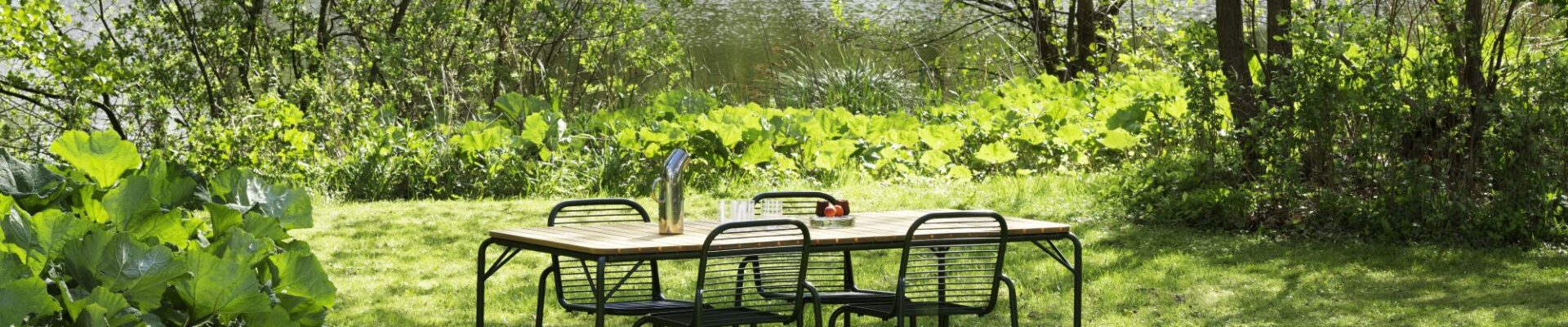 A vibrant outdoor scene features a wooden rectangular dining table with four black metal chairs, set on a lush green lawn.