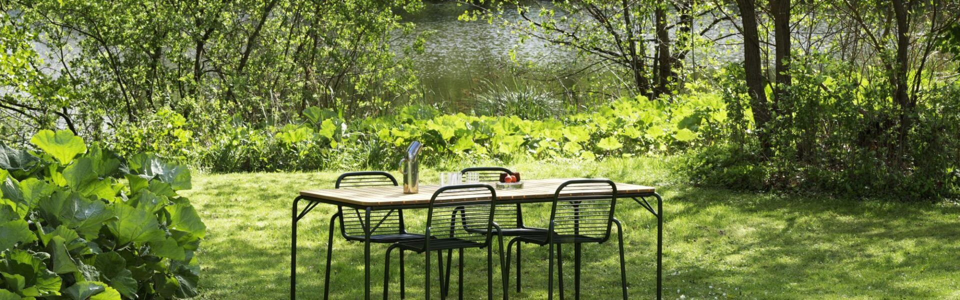 A vibrant outdoor scene features a wooden rectangular dining table with four black metal chairs, set on a lush green lawn.