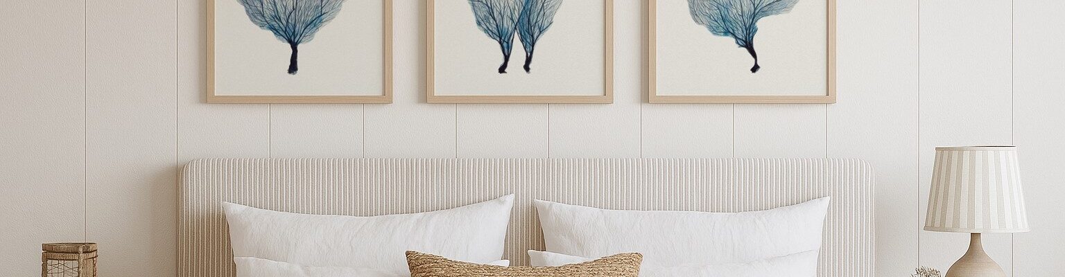 A bedroom scene features a large bed with a white pleated headboard and white bedding. A textured brown throw pillow rests on the bed. Above the bed, a set of three framed art prints, each depicting blue coral, are hung on a white panelled wall.