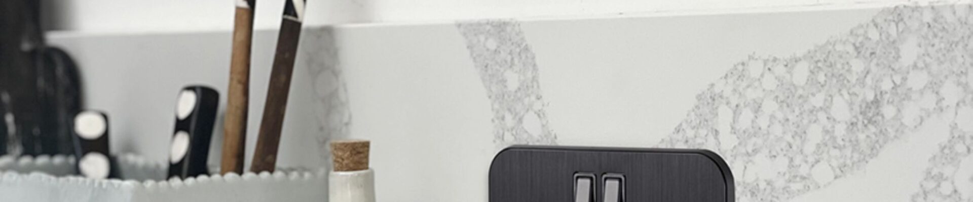 A sleek, black double UK plug socket (13A) is mounted on a grey and white marble-effect splashback in a modern kitchen.