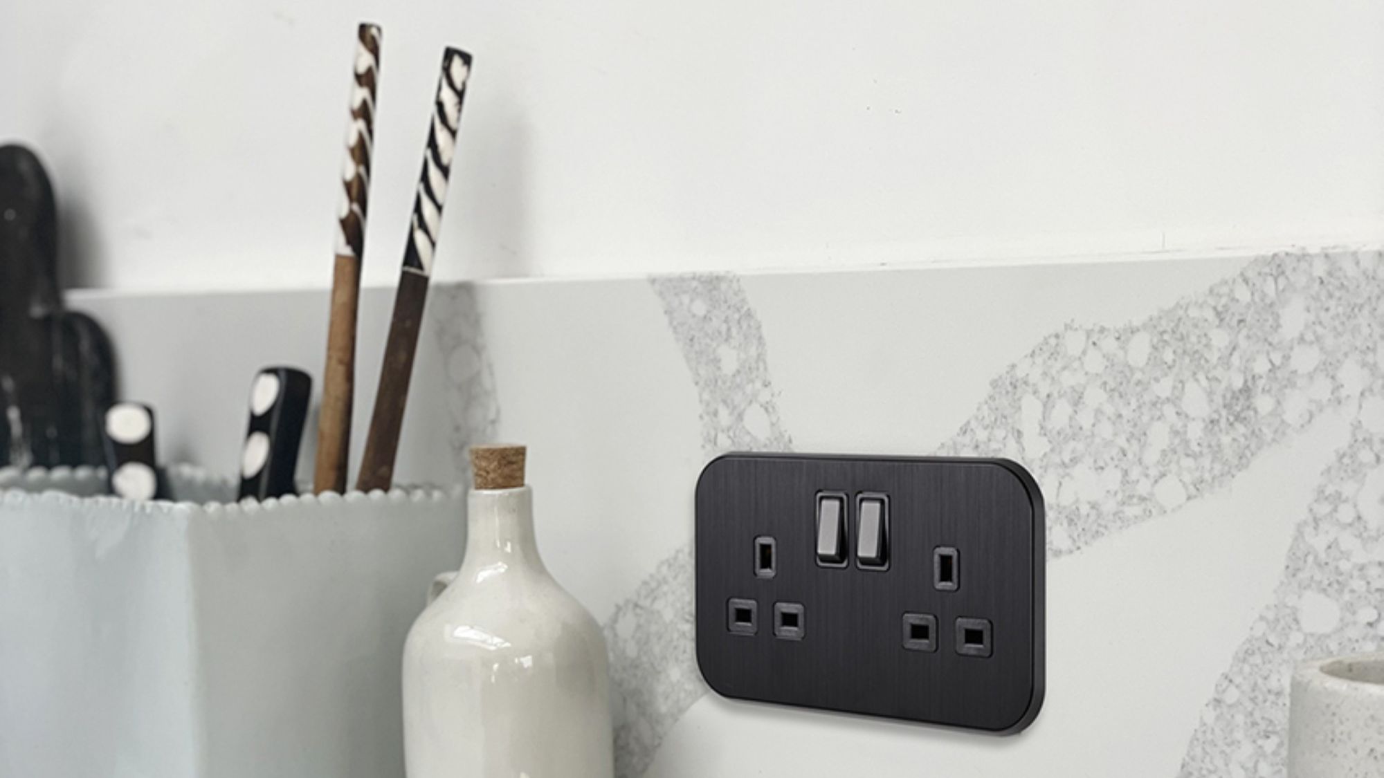 A sleek, black double UK plug socket (13A) is mounted on a grey and white marble-effect splashback in a modern kitchen.