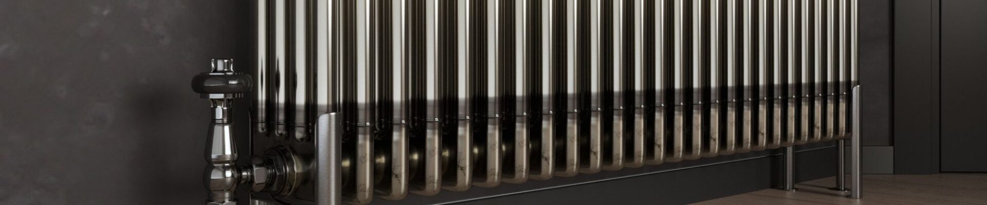 A bare metal, multi-column radiator stands on a light wooden floor with dark grey skirting boards and a dark grey wall behind it. To the left, a silver valve and pipe connect to the radiator, and on the right, two matching silver feet support it.