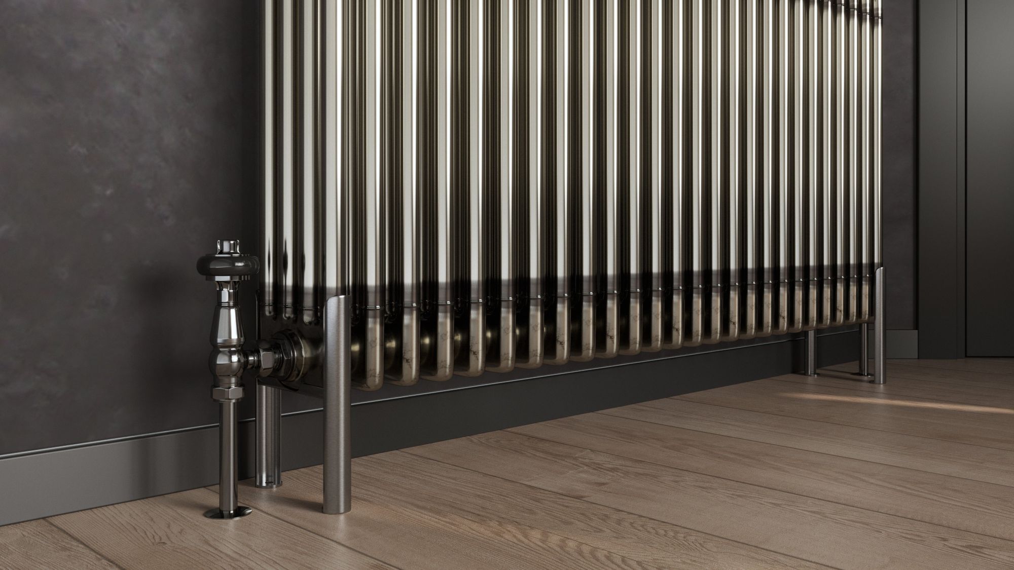 A bare metal, multi-column radiator stands on a light wooden floor with dark grey skirting boards and a dark grey wall behind it. To the left, a silver valve and pipe connect to the radiator, and on the right, two matching silver feet support it.