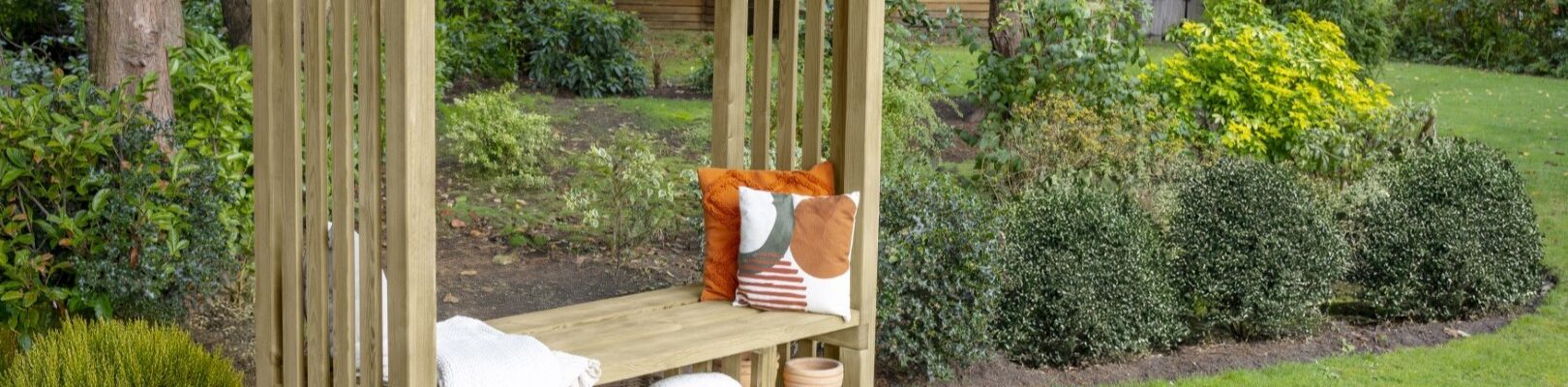 A natural wood-stained pergola with an integrated bench seat is positioned on a vibrant green lawn. The pergola has a rectangular shape with slatted sides and an open roof, from which hangs a string of decorative globe lights. On the bench, there are several cushions in warm tones of orange, brown, and cream.