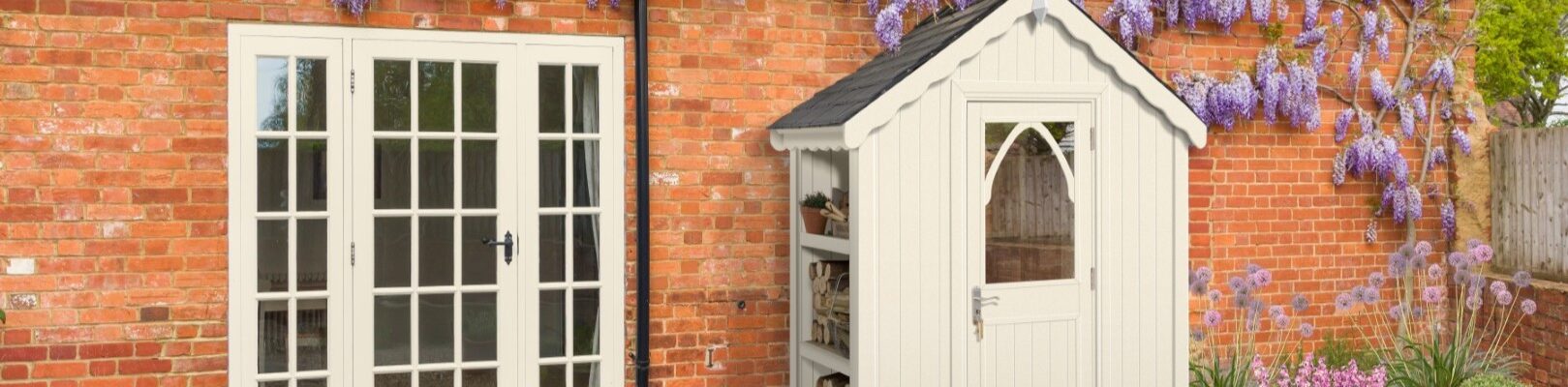 A quaint cream-coloured shed stands nestled beside a rustic brick house. The shed features a dark roof, a charming arched window in its door, and an open shelving unit on its left side, holding what appears to be firewood and a potted plant.
