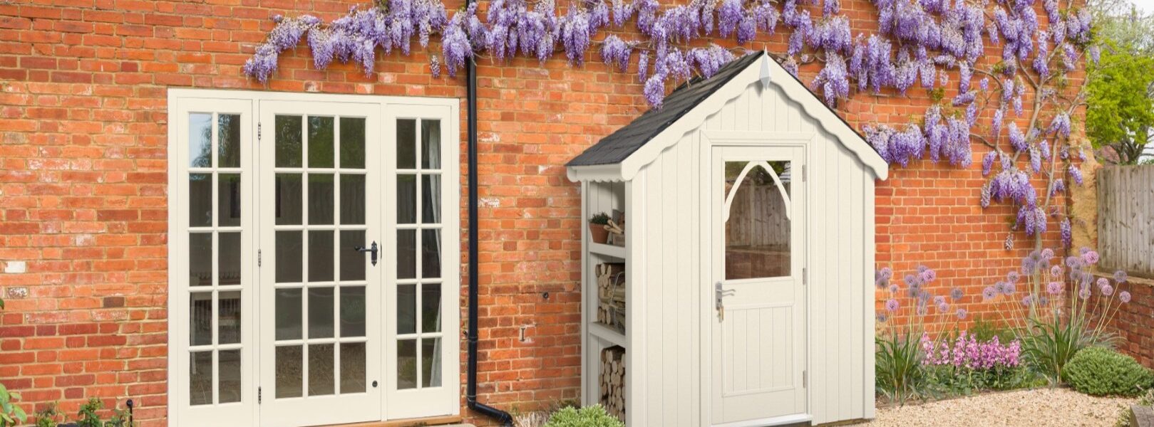 A quaint cream-coloured shed stands nestled beside a rustic brick house. The shed features a dark roof, a charming arched window in its door, and an open shelving unit on its left side, holding what appears to be firewood and a potted plant.