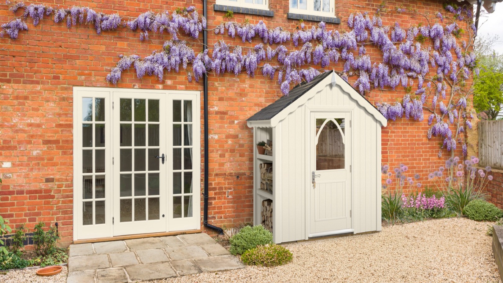 A quaint cream-coloured shed stands nestled beside a rustic brick house. The shed features a dark roof, a charming arched window in its door, and an open shelving unit on its left side, holding what appears to be firewood and a potted plant.
