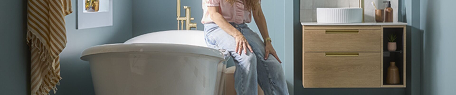 A woman with long, dark hair is seated on the edge of a white, freestanding bathtub in a bathroom. She is wearing a light-coloured, long-sleeved top and blue jeans. To her right, there is a wooden wall-mounted vanity unit with a white circular sink and a gold-coloured faucet.